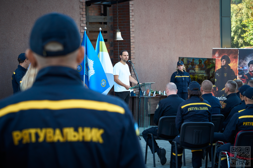 Олександр Коваль стоїть за прозорою трибуною та звертається до рятувальників, які сидять на кріслах та слухають його