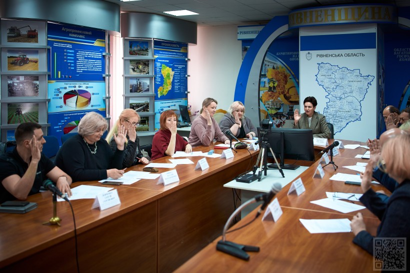 У залі за столом сидить група людей на чолі із застукпницею голови Рівненської ОДА Людмилою Шактовською. Перед ними таблички та документи. Вони підняли руку та голосують.