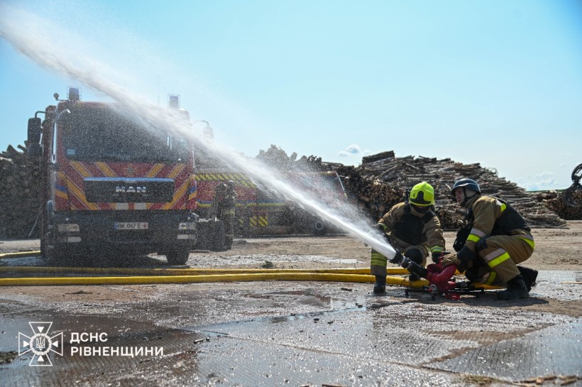На фото мамент навчання: пожежний автомобіль на задньому плані, а попереду двоє рятувальників поливають водою предмет, що поза кадром