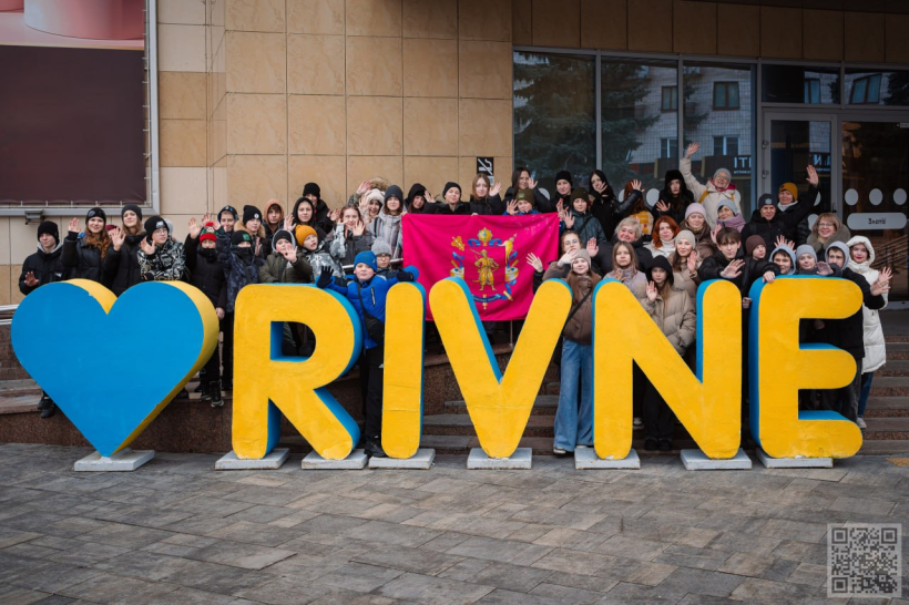 На фото зображена велика група дітей та підлітків разом із кількома дорослими, які стоять на сходах біля сучасної будівлі. Усі одягнені по-зимовому — у куртки, шапки, шарфи, що свідчить про прохолодну пору року. Більшість учасників усміхаються та махають руками в камеру, створюючи атмосферу дружності й позитиву.

У центрі групи розгорнуто малиновий прапор із гербом. Попереду встановлена велика декоративна інсталяція з написом «RIVNE»: синє серце та жовті літери, що символізують любов до міста і виконані в кольорах українського прапора.

Світлина передає відчуття єдності, гордості за рідне місто та спільності. Атмосфера тепла й піднесена, попри холодну погоду.