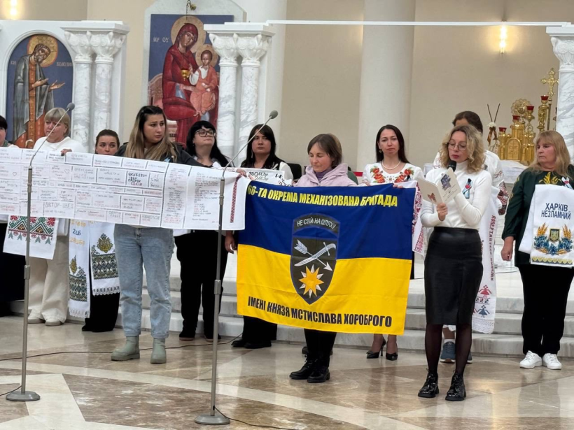 У просторому світлому залі стоїть група жінок, які тримають у руках жовто-блакитний прапор на якому герб та напис: 66-та окрема механізована бригада. Ще одна жінка тримає у руках вишитий рушник
