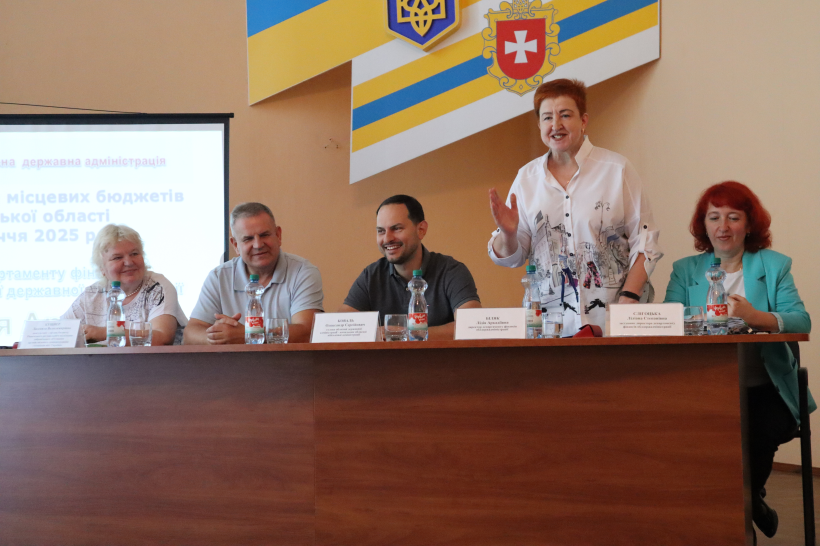 У президії сидять: начальник Рівненської ОВА Олександр Коваль та інші спікери семінару-навчання. Директор Департаменту фінансів ОДА Лідія Біляк стоїть біля столу і звертається до присутніх жестикулюючи руками, начальник ОВА посміхається.