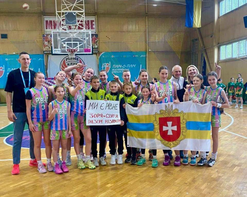 Рівненська дівчача команда з баскетболу стоять у спортзалі. Вони посміхаються та тримають у руках прапор Рівного