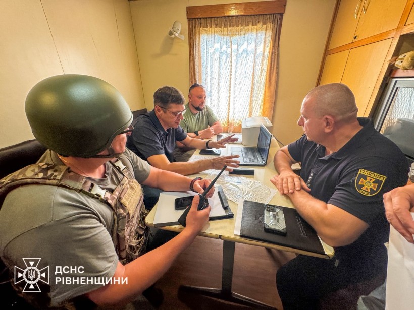 За столдом сидять члени штабу із ліквідації наслідків аварії. Серед них перший заступник голови Рівненської ОДА Сергій Подолін. Вони сидять за столом і дивляться у монітор ноутбуку, один із учасників у військовій формі, бронежилеті та із рацією у руках