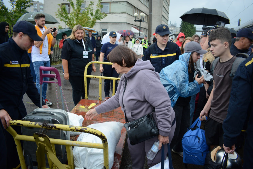 На платформі вокзалу міста Рівне жінка із багажного візка бере свою сумку. Поруч багато людей: представниці ДСНС, Центру життєстійкості та інших громадських та державних установ та організацій. Вони чекають щоб допомогти вимушеним переселенцям та зайнятися їхнім поселенням