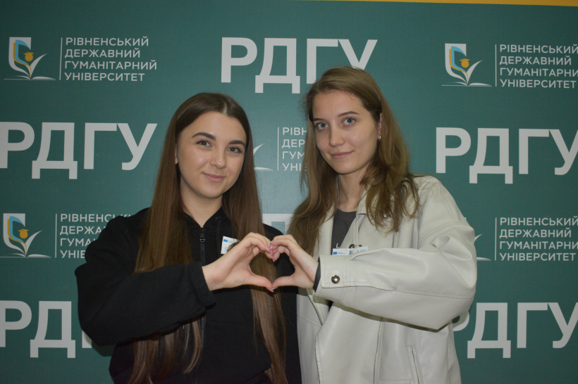 Двоє студенток стоять біля банера із написом "РДГУ" та склали з рук сердечко