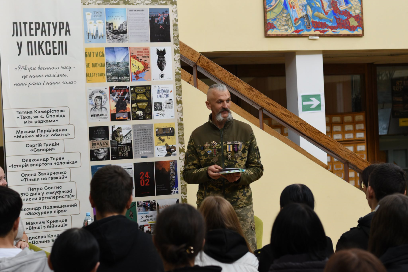 Чоловік у формі ЗСУ із книгою в руках, колоритними козацькими вусами та орденами на грудях виступає перед учнями старших класів у бібліотеці. Молодь уважно слухає ветерана.