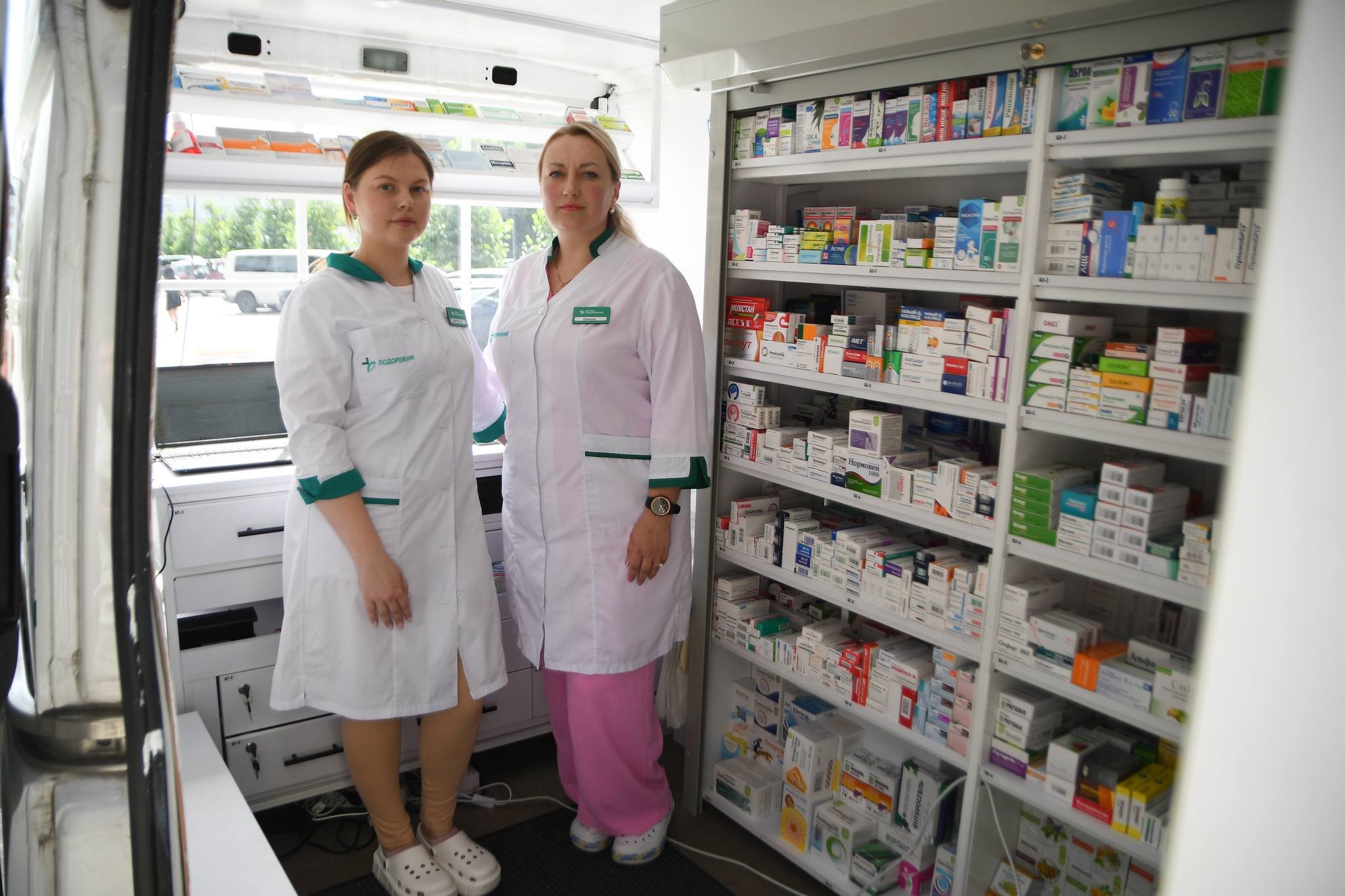 Дві фармацевтки в мобільній аптеці на фоні стелажу з медичнимим препаратами