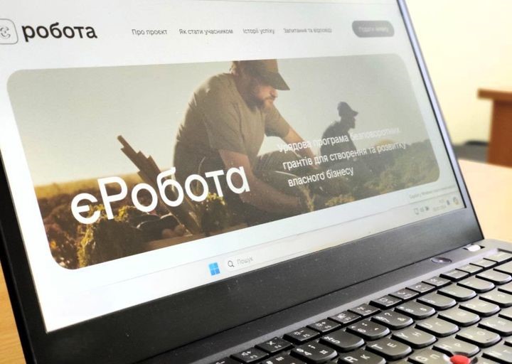 Екран ноутбука, на якому відкритий вебсайт. Вебсайт має назву "єРобота" і містить інформацію про урядову програму грантів для створення та розвитку власного бізнесу. На банері зображена людина, ймовірно фермер, який працює на полі.

На верхній панелі вебсайту розміщені наступні вкладки:
Про проект
Як стати учасником
Історії успіху
Запитання та відповіді