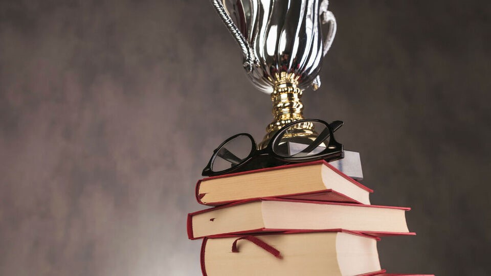 Стопка книг, на верхівці якої розміщені окуляри та чаша переможця. Книги мають червоні корінці, і зверху на них лежать чорні окуляри. Чаша переможця розташована на самій вершині стопки книг. Фон зображення має сіро-коричневий відтінок.