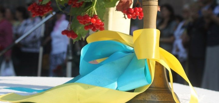 Традиційний шкільний дзвінок, перев’язаний жовто-блакитною стрічкою, що символізує український прапор. Біля дзвінка розташована гілка калини з яскраво-червоними ягодами.